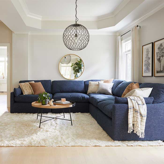 A dark blue fabric sectional with neutral throw pillows in a white-walled room. A black globe chandelier is overhead and a plush beige rug is underneath.