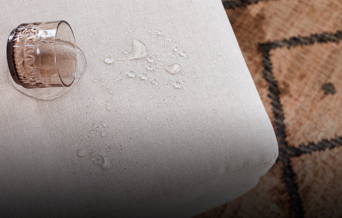 Tinted drinking glass of spilled water on a beige fabric ottoman on a tan rug