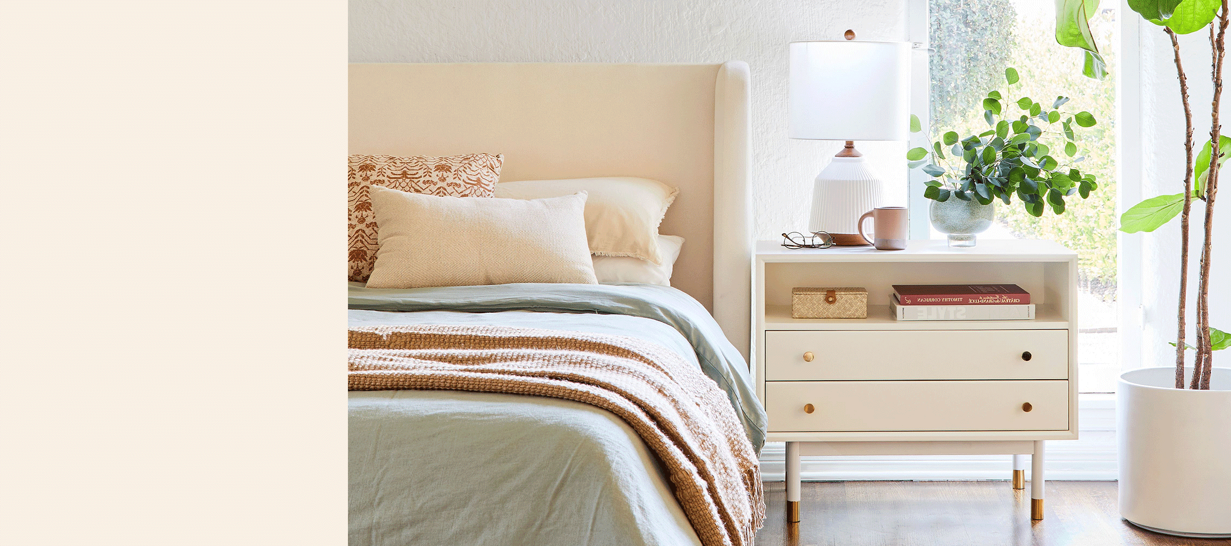 A beige upholstered headboard with beige and rust throw pillows, a clay colored blanket, a sage green duvet cover. At the right, a wide white and brass nightstand with two drawers. Atop, a white and wood table lamp and a vase with greener.